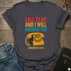 Gray t-shirt with vintage phone graphic and text Call to Me and I Will Answer You Jeremiah 33:3 retro phone faith Christian apparel conversation starter on a wooden surface with jeans and a Bible.