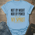 A light blue graphic t-shirt laid flat on a rustic wooden surface, styled with blue jeans and a black leather Bible. The t-shirt features the text 'NOT BY MIGHT NOR BY POWER BUT BY MY SPIRIT ZECHARIAH 4:6' in black and mustard yellow lettering.