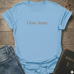 A light blue crewneck t-shirt with the text 'I love Jesus' printed in brown across the chest, styled in a flat-lay arrangement with blue jeans and a Bible on a rustic wooden background.