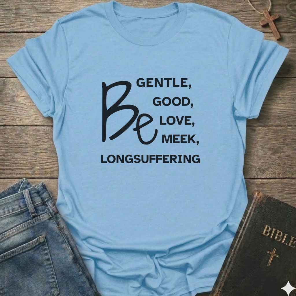 A light blue graphic t-shirt laid flat on a rustic wooden background, styled with a pair of blue jeans and a dark leather Bible. The t-shirt features black text that reads 'BE GENTLE, GOOD, LOVE, MEEK, LONGSUFFERING' with 'Be' written in a large script font.