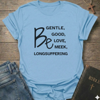 A light blue graphic t-shirt laid flat on a rustic wooden background, styled with a pair of blue jeans and a dark leather Bible. The t-shirt features black text that reads 'BE GENTLE, GOOD, LOVE, MEEK, LONGSUFFERING' with 'Be' written in a large script font.