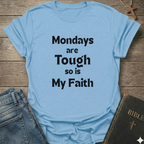 A light blue graphic t-shirt with the text 'Mondays are Tough so is My Faith' printed in black. The t-shirt is styled in a flat-lay arrangement on a rustic wooden background, accompanied by a pair of blue jeans and a dark leather Bible with a gold cross.