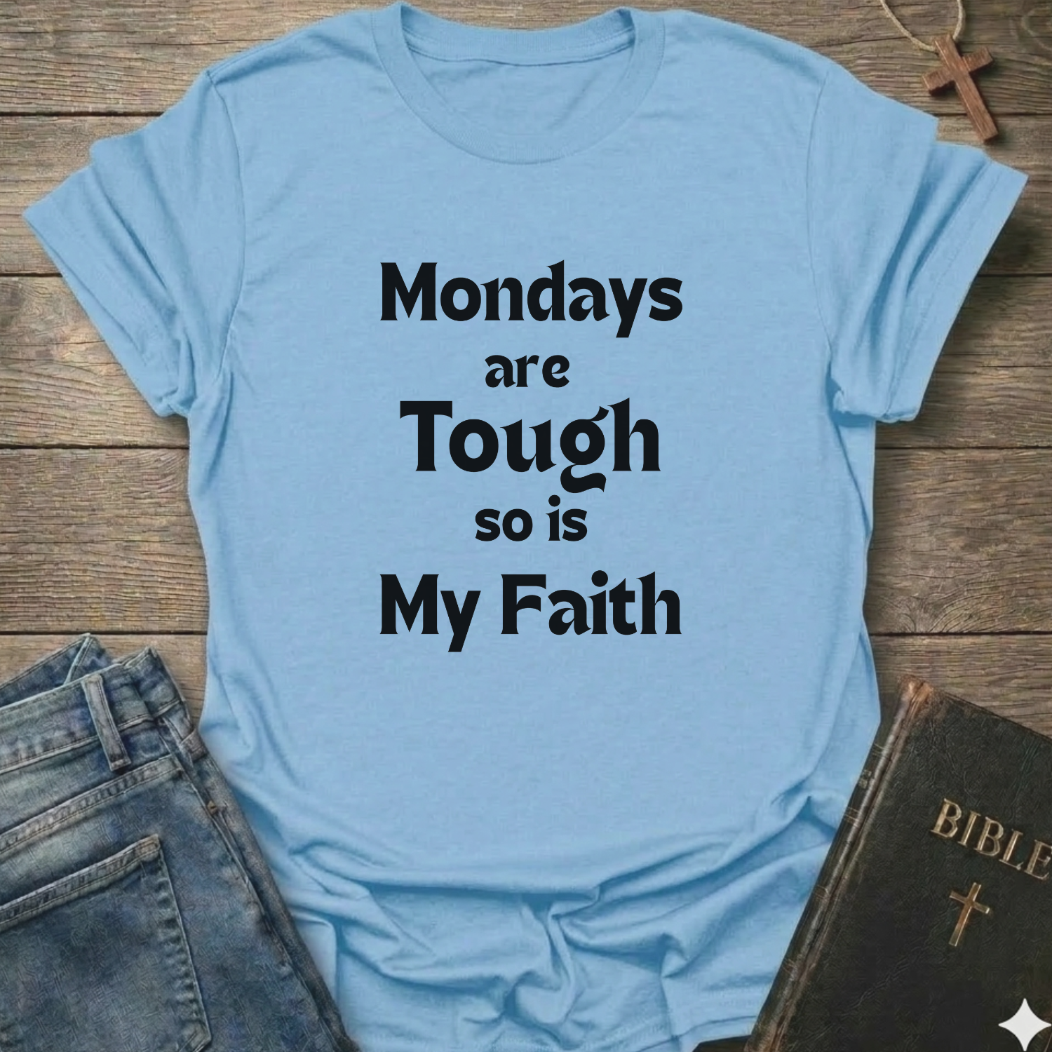 A light blue graphic t-shirt with the text 'Mondays are Tough so is My Faith' printed in black. The t-shirt is styled in a flat-lay arrangement on a rustic wooden background, accompanied by a pair of blue jeans and a dark leather Bible with a gold cross.