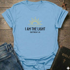 A light blue graphic t-shirt laid flat on a rustic wooden background, styled with a pair of blue jeans and a black Bible. The t-shirt features a yellow sun graphic and the text 'I AM THE LIGHT MATTHEW 5:14' printed on the chest.