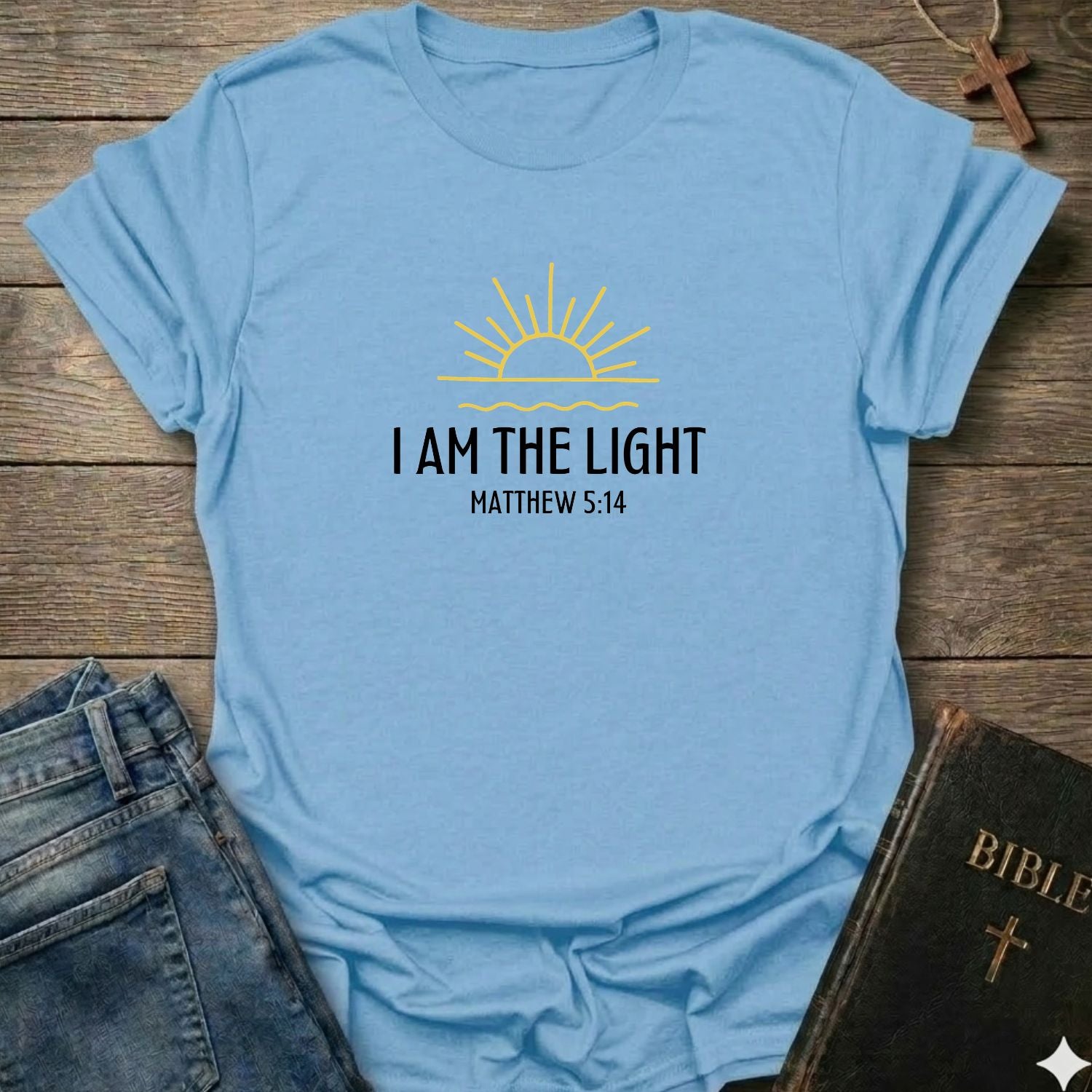A light blue graphic t-shirt laid flat on a rustic wooden background, styled with a pair of blue jeans and a black Bible. The t-shirt features a yellow sun graphic and the text 'I AM THE LIGHT MATTHEW 5:14' printed on the chest.