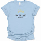 A light blue short-sleeve t-shirt with a graphic on the chest featuring a yellow sun and the text 'I AM THE LIGHT MATTHEW 5:14' in black. The shirt is displayed flat against a clean white background.
