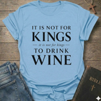 A light blue graphic t-shirt with white text reading 'IT IS NOT FOR KINGS it is not for kings TO DRINK WINE', styled in a flat-lay on a rustic wooden background with blue jeans and a Bible nearby.