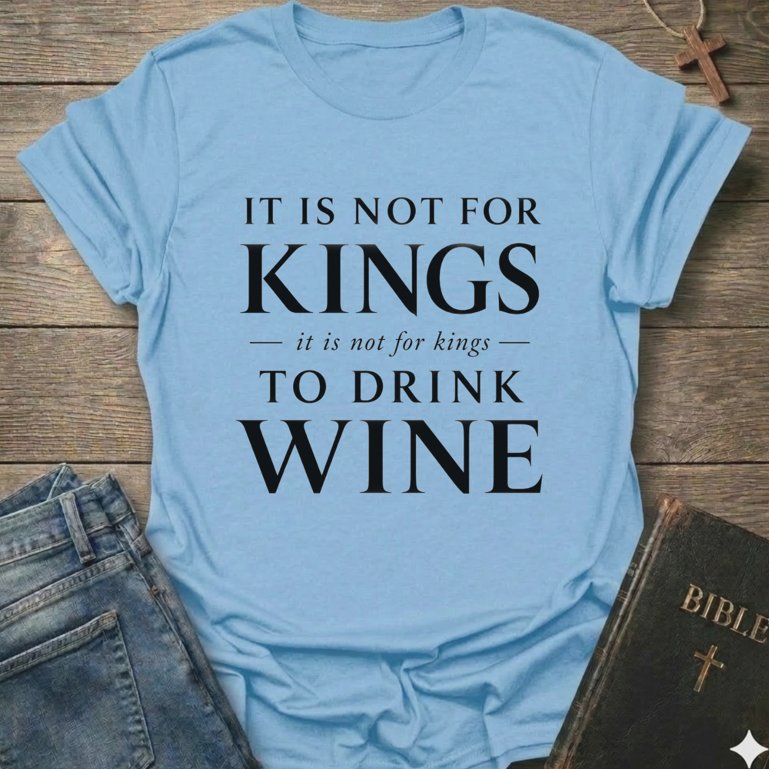 A light blue graphic t-shirt with white text reading 'IT IS NOT FOR KINGS it is not for kings TO DRINK WINE', styled in a flat-lay on a rustic wooden background with blue jeans and a Bible nearby.
