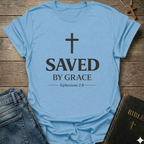 Light blue Christian t-shirt with 'Saved by Grace' text and cross reference Ephesians 2:8 scripture faith apparel conversation starter  on a wooden surface with jeans and a Bible. 