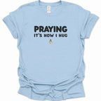 A light blue short-sleeve t-shirt with a graphic print that reads 'PRAYING IT'S HOW I HUG' in black text, with a small praying hands illustration below. The shirt is displayed flat against a clean white background.