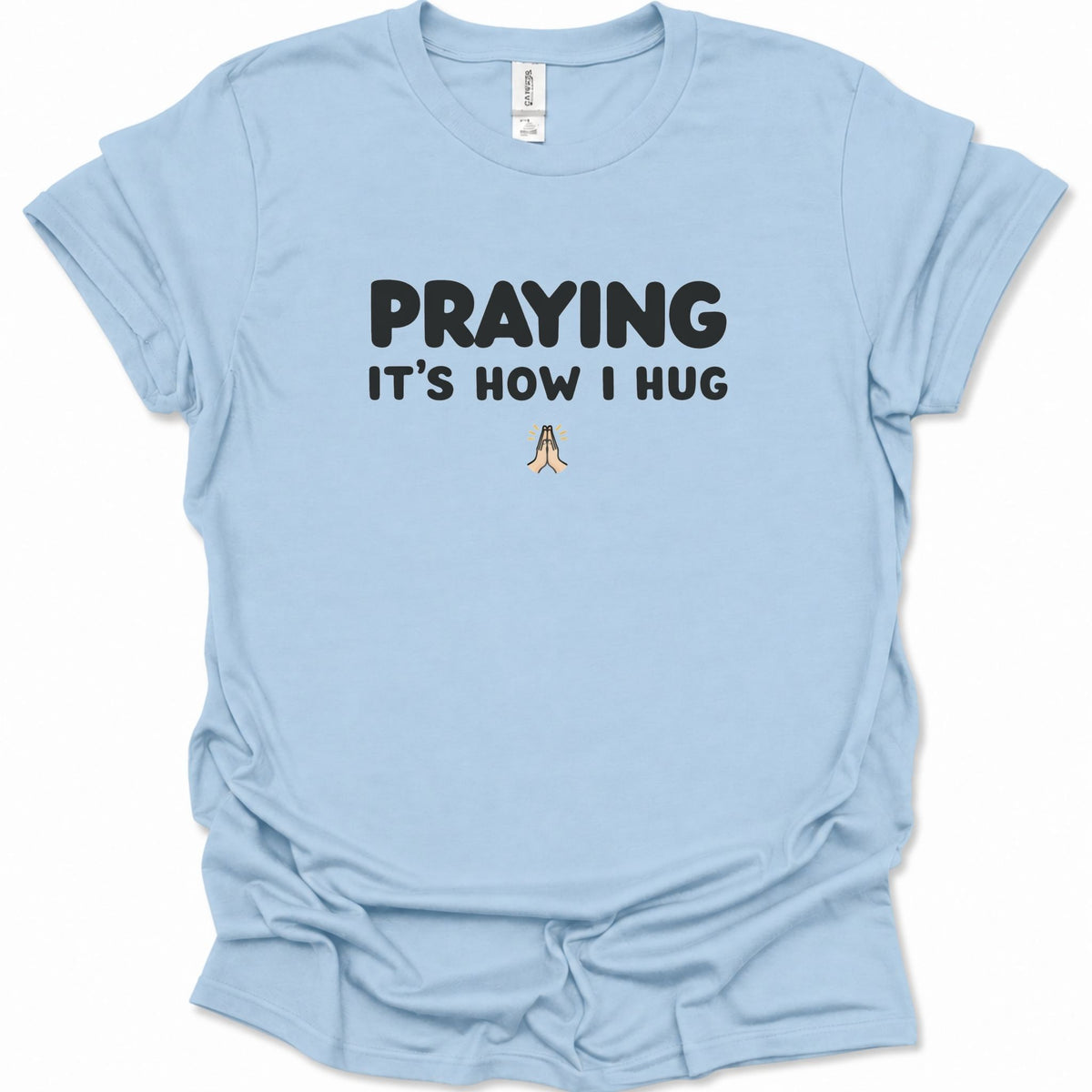 A light blue short-sleeve t-shirt with a graphic print that reads 'PRAYING IT'S HOW I HUG' in black text, with a small praying hands illustration below. The shirt is displayed flat against a clean white background.