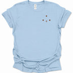 A light blue short-sleeve crewneck t-shirt with a small minimalist graphic on the left chest consisting of three brown dots and a brown cross, displayed on a clean white background.