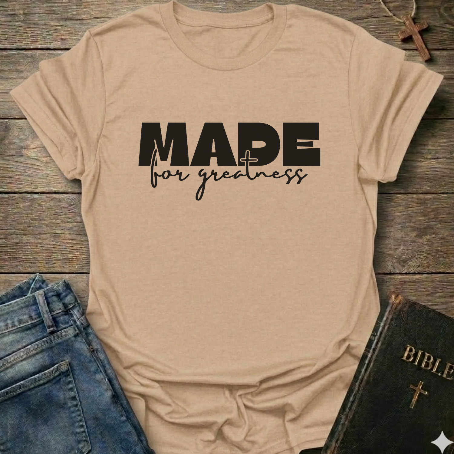 A natural short sleeve T-shirt laid flat on a rustic wooden surface, with the text 'MADE for greatness' based on 2 Corinthians 5 verse 17 printed on the front, styled in a flat-lay arrangement with blue jeans and a Bible on a rustic wooden background.