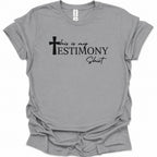 A sport gray short-sleeve t-shirt with a black graphic text print reading 'this is my TESTIMONY Shirt' accompanied by a cross icon, displayed on a clean white background.