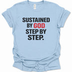 A light blue short-sleeve t-shirt with the text 'SUSTAINED BY GOD STEP BY STEP.' printed on the front in black and red lettering, displayed flat against a white background.