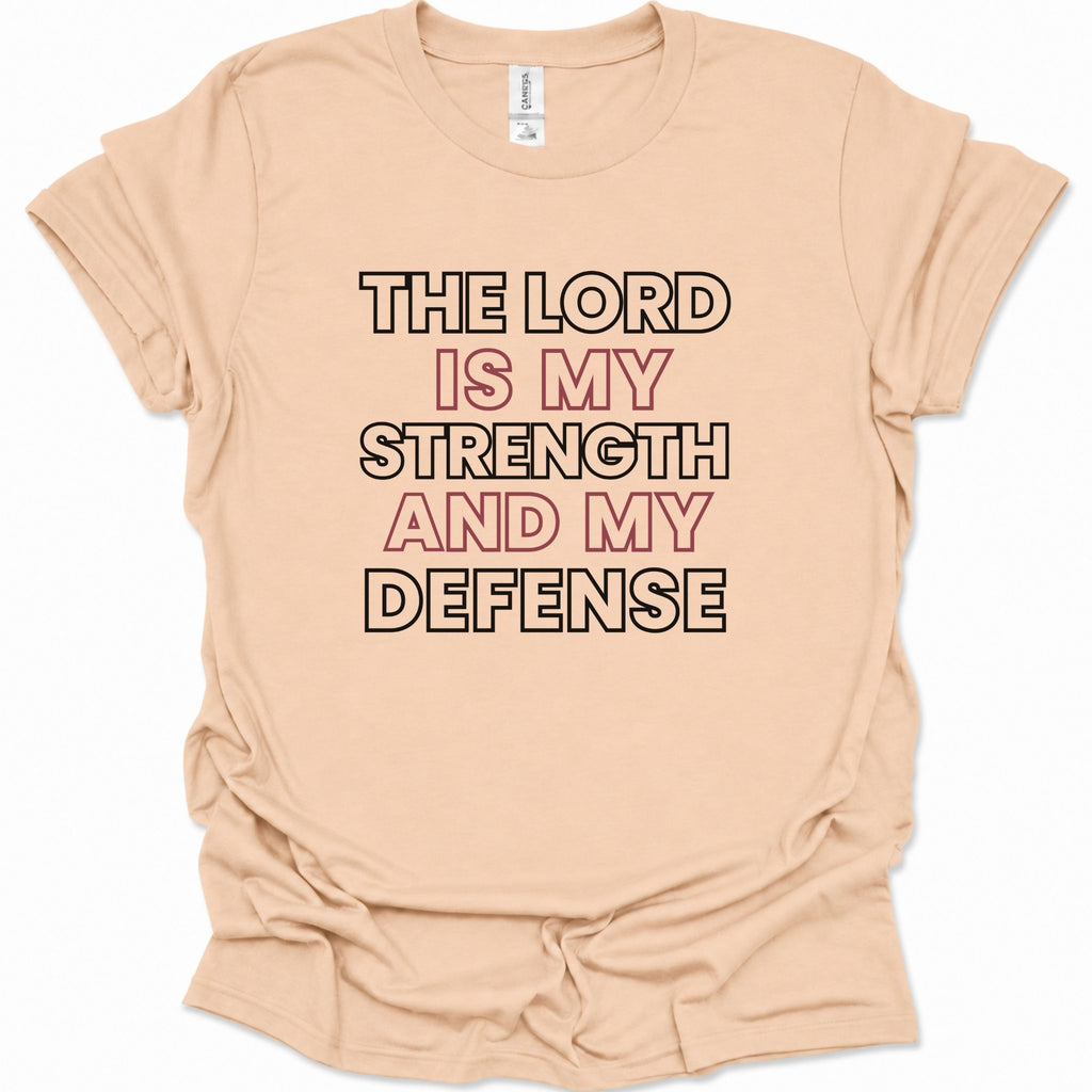 A light natural-colored short-sleeve t-shirt with a graphic text print reading 'THE LORD IS MY STRENGTH AND MY DEFENSE' in black and dusty pink lettering, laid flat against a white background.