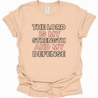 A light natural-colored short-sleeve t-shirt with a graphic text print reading 'THE LORD IS MY STRENGTH AND MY DEFENSE' in black and dusty pink lettering, laid flat against a white background.