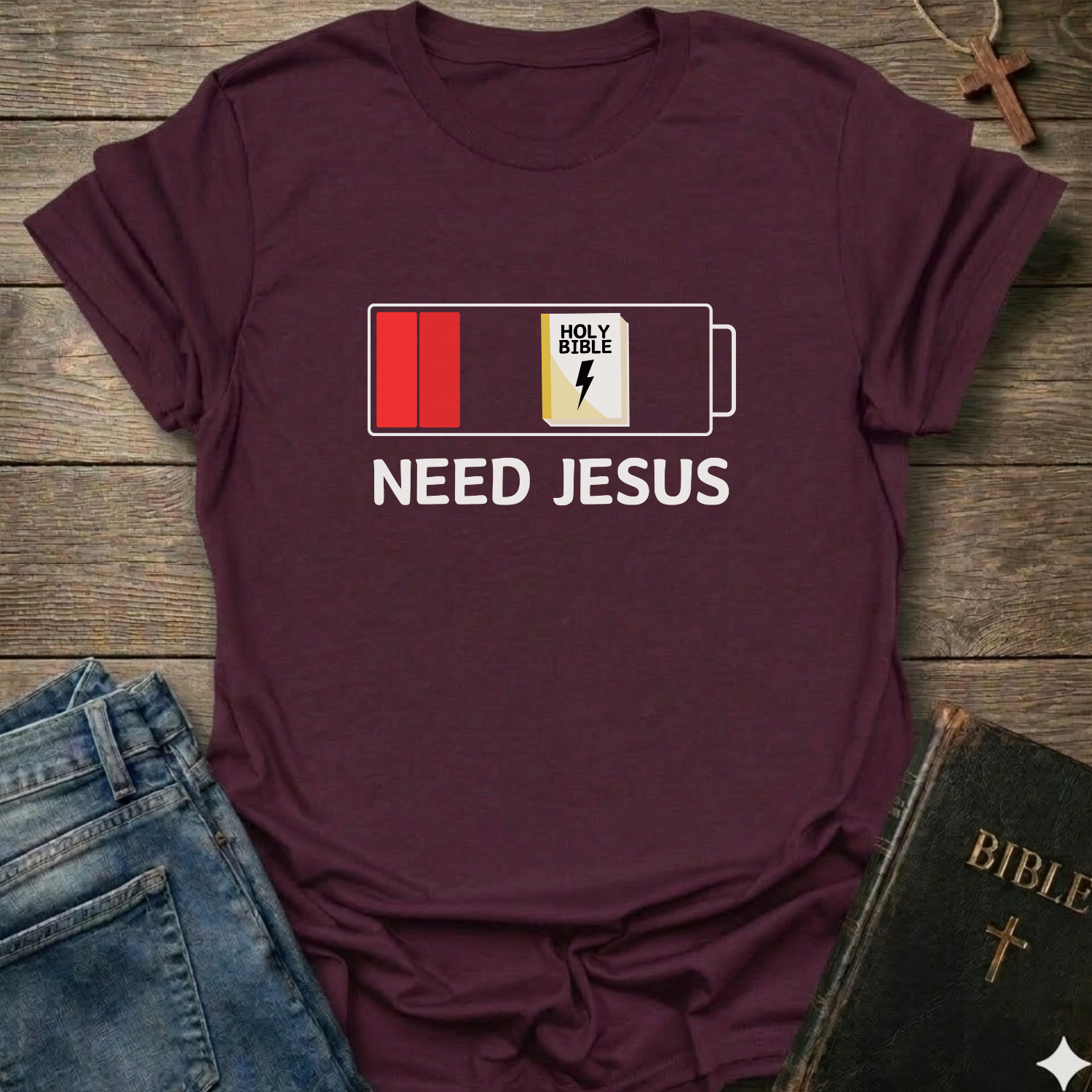 Maroon t-shirt with 'Need Jesus' graphic on a wooden surface with jeans and a Bible. Need Jesus to Recharge My Battery Christian T-Shirt bold faith apparel Faith Forward