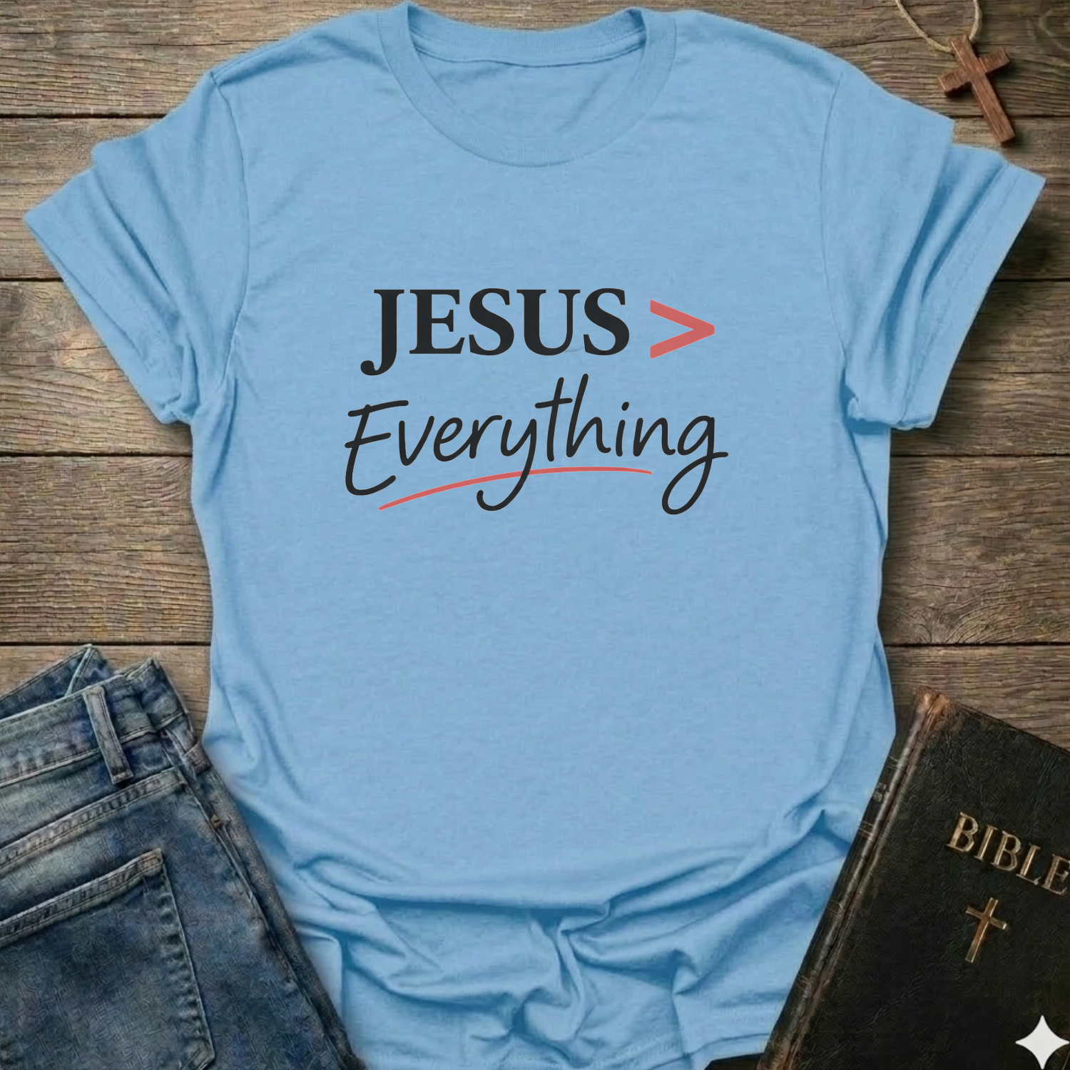 Light blue T-Shirt faith apparel conversation starter with 'JESUS > Everything' text on a wooden surface with jeans and a Bible.