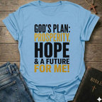 A light blue graphic t-shirt laid flat on a rustic wooden background, styled with a pair of blue jeans and a black Bible. The t-shirt features the text 'GOD'S PLAN: PROSPERITY, HOPE & A FUTURE FOR ME!' in black and mustard yellow lettering.