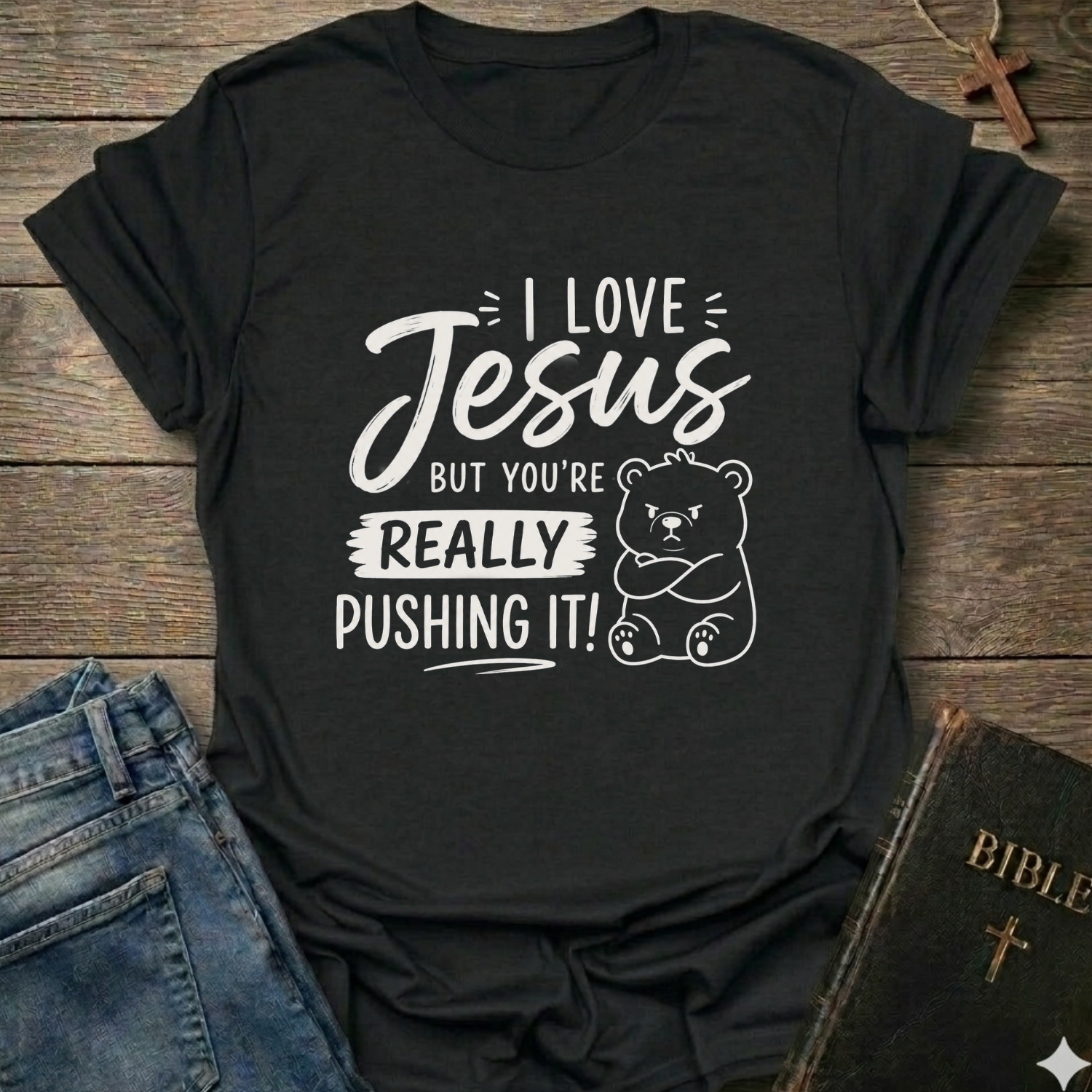 A black short-sleeve graphic T-shirt laid flat on a rustic wooden surface, styled with blue jeans, a Bible, and a small cross. The shirt has white text reading 'I Love Jesus But You're Really Pushing It!' plus a white teddy bear illustration.