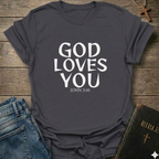 Heather Grey t-shirt with 'GOD LOVES YOU' John 3:16 text Christian bold faith apparel Faith Forward on a wooden surface with jeans and a Bible.