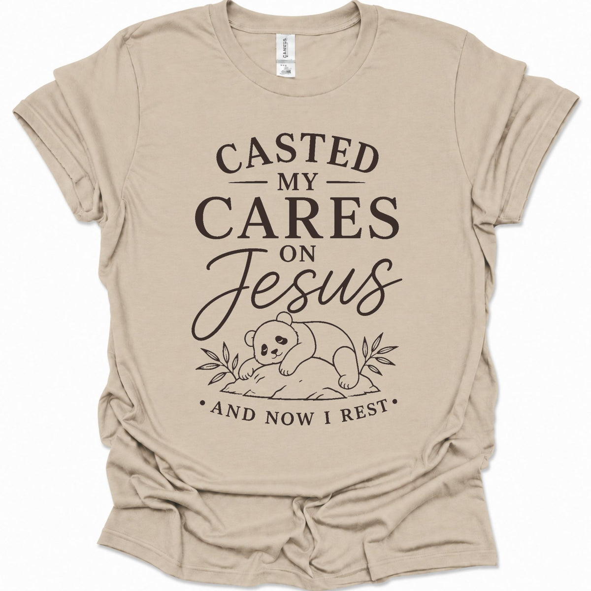 A sand colored short-sleeve t-shirt with a black graphic print that reads 'CASTED MY CARES ON Jesus AND NOW I REST' with a cartoon bear illustration, shown on a plain white background.