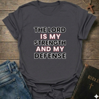 Gray t-shirt with 'The Lord is my strength and my defense' Exodus 15:2 bold faith apparel Faith Forward on a wooden surface with jeans and a Bible.