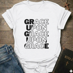White t-shirt with 'Grace Upon Grace Upon Grace' Christian T-Shirt John 1:16 bold faith apparel text on a wooden surface with jeans and a Bible.