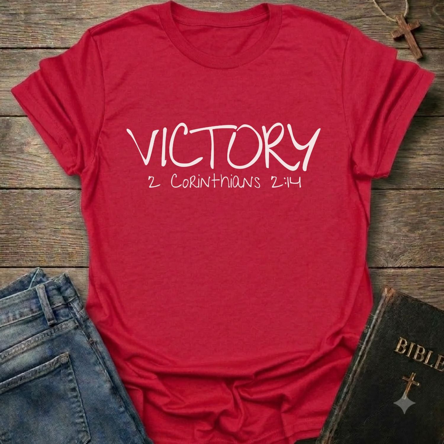 A red short-sleeve t-shirt with 'VICTORY 2 CORINTHIANS 2:14' printed in white on the front, styled in a flat lay with blue jeans and a Bible on a rustic wooden background.