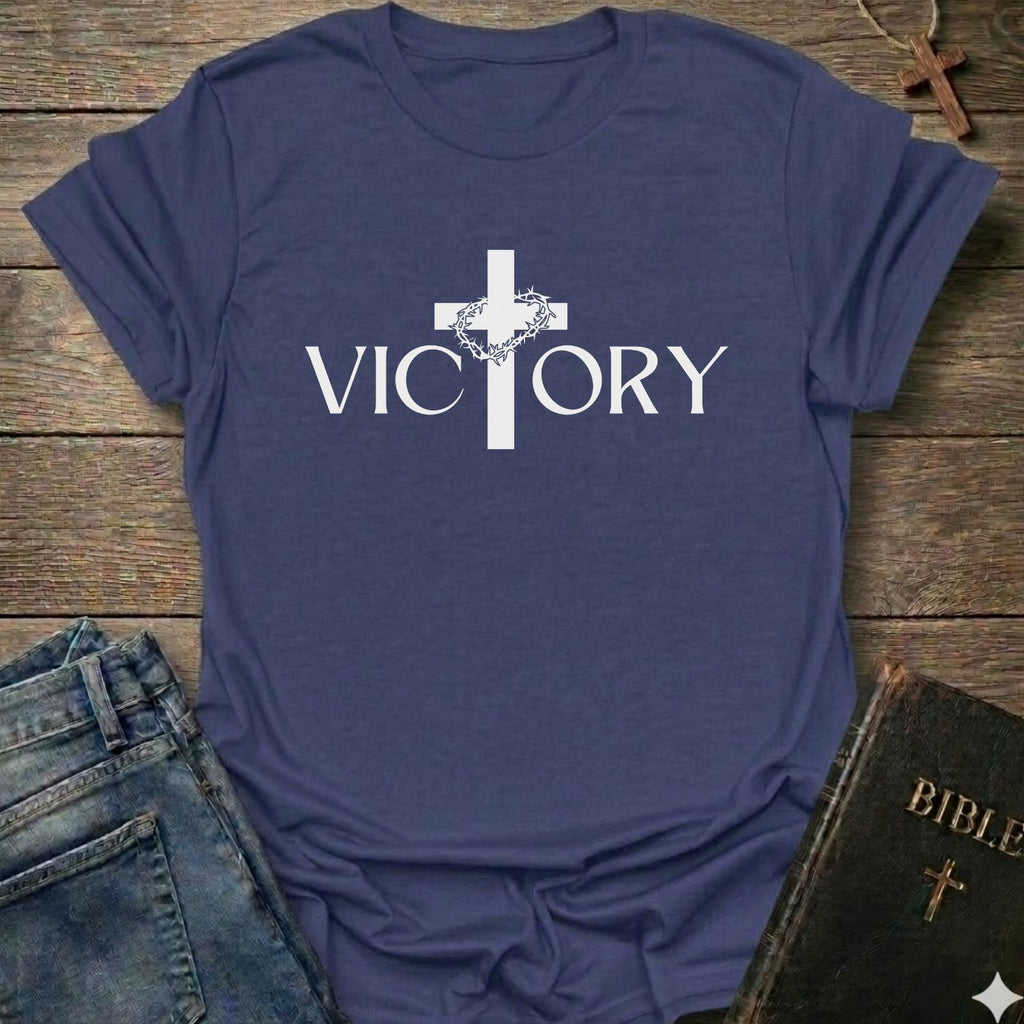 A purple blue short-sleeve t-shirt with a white graphic print of a cross and the word 'VICTORY' on the front, styled in a flat-lay arrangement with jeans, a Bible, and a wooden cross on a rustic wood background.