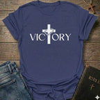 A purple blue short-sleeve t-shirt with a white graphic print of a cross and the word 'VICTORY' on the front, styled in a flat-lay arrangement with jeans, a Bible, and a wooden cross on a rustic wood background.