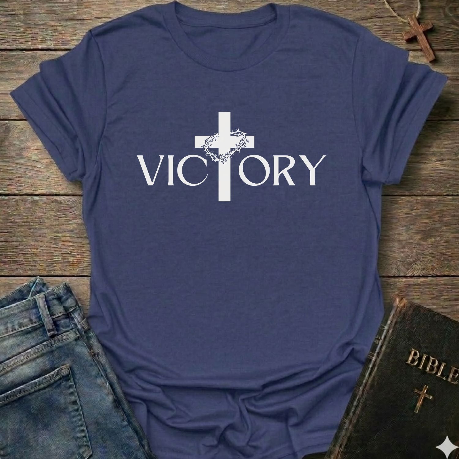 A purple blue short-sleeve t-shirt with a white graphic print of a cross and the word 'VICTORY' on the front, styled in a flat-lay arrangement with jeans, a Bible, and a wooden cross on a rustic wood background.