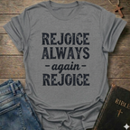 Gray t-shirt with 'REJOICE ALWAYS again REJOICE' text Philippians 4:4 bold faith apparel Faith Forward on a wooden surface with jeans and a Bible.