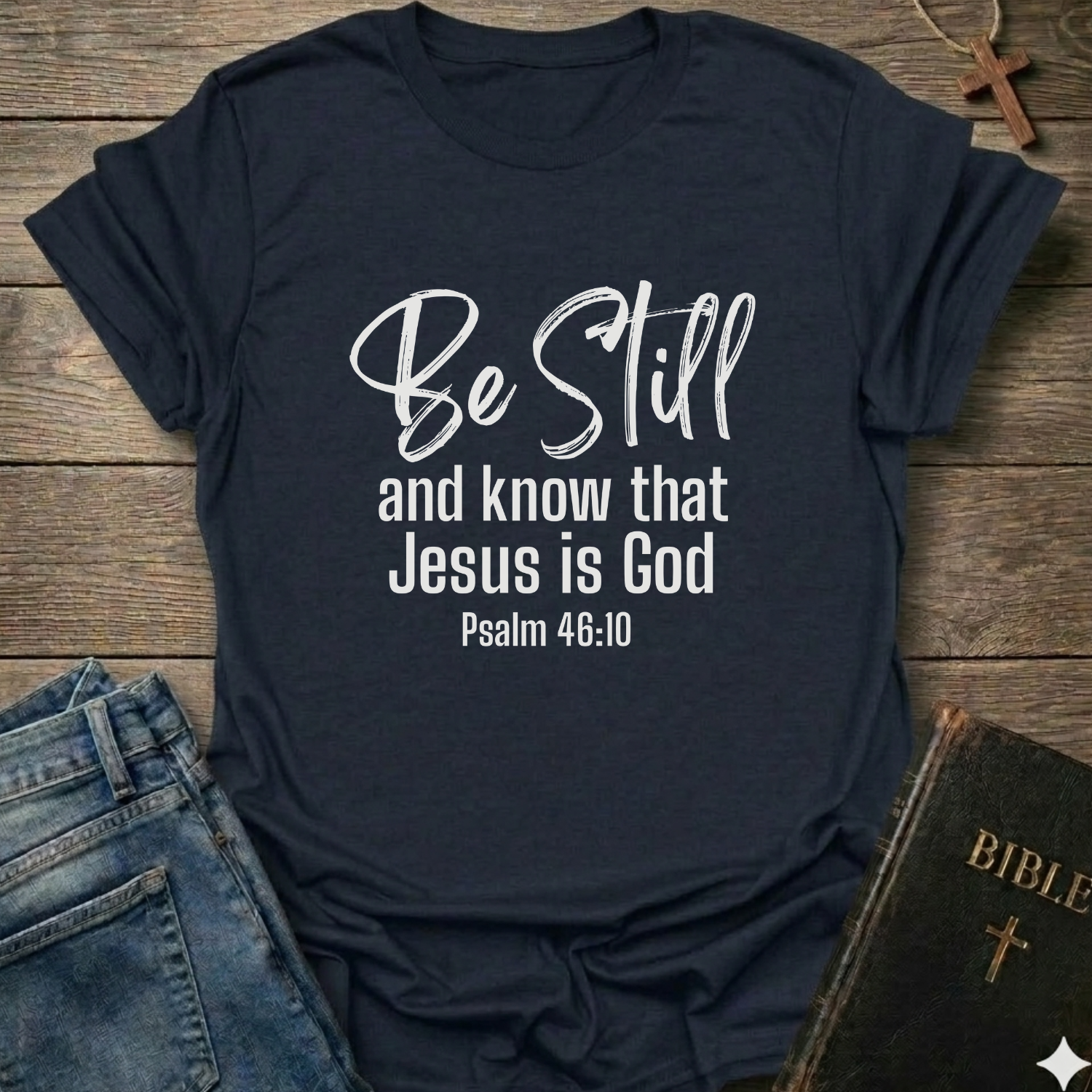 Navy blue t-shirt with 'Be Still and know that Jesus is God' text Psalm 46:10 script faith apparel Faith Forward on a wooden surface with jeans and a Bible.