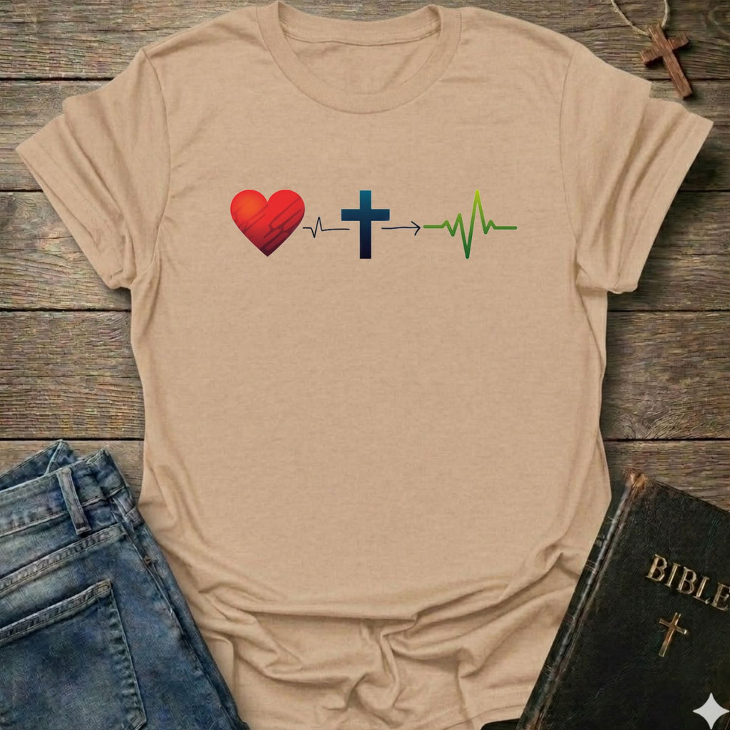 A natural graphic t-shirt laid flat on a rustic wooden background, styled with blue jeans and a black Bible. The t-shirt features a graphic of a red heart, a blue Christian cross, and a green heartbeat line connected by an arrow.