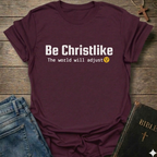 Maroon t-shirt with 'Be Christlike The world will adjust' text on a wooden surface with jeans and a Bible.