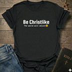 A black short-sleeve graphic T-shirt laid flat on a rustic wooden surface, styled with blue jeans, a Bible, and a cross. The shirt has white text reading 'Be Christlike' with 'The world will adjust' and a small yellow smiley face underneath.