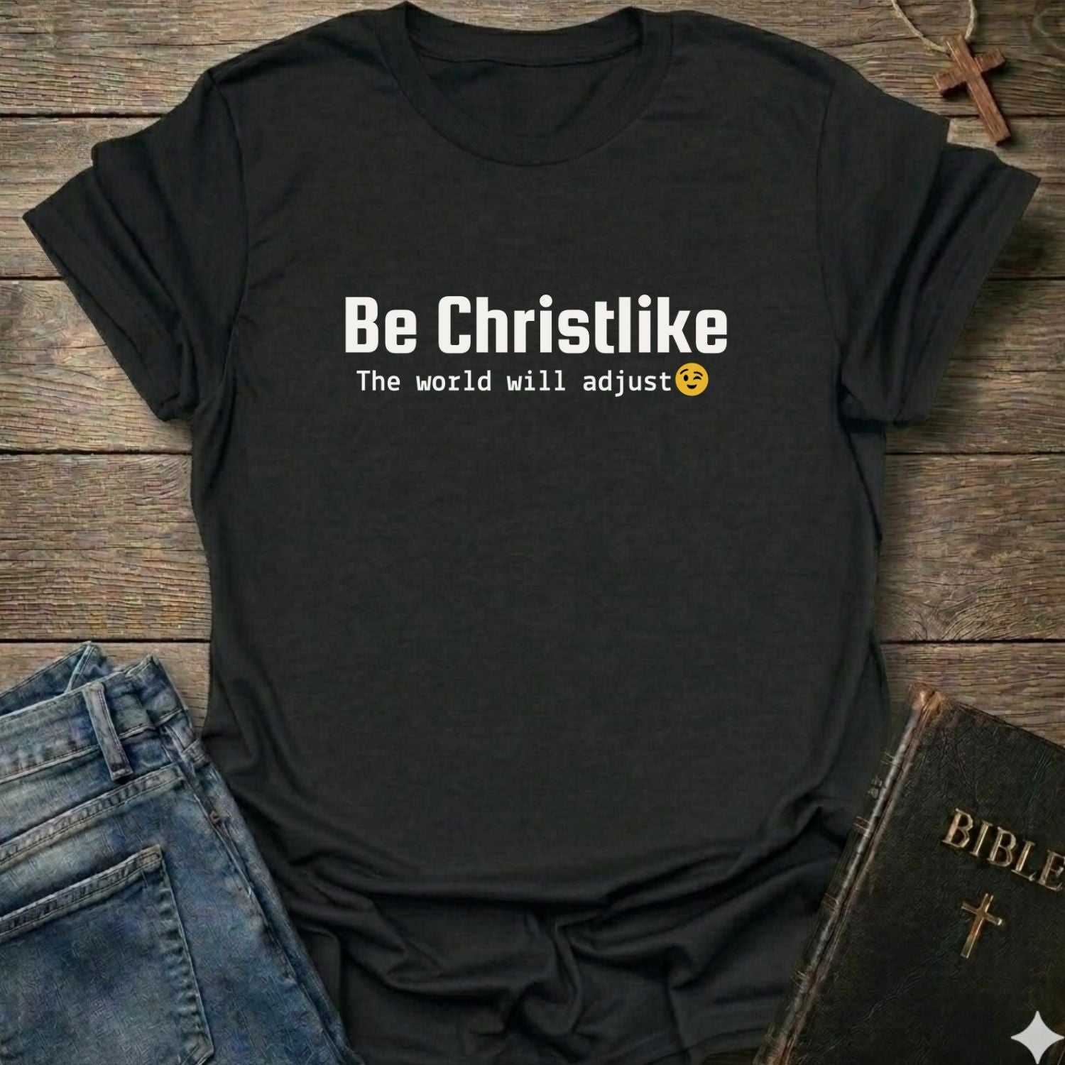A black short-sleeve graphic T-shirt laid flat on a rustic wooden surface, styled with blue jeans, a Bible, and a cross. The shirt has white text reading 'Be Christlike' with 'The world will adjust' and a small yellow smiley face underneath.