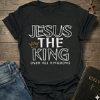 Black t-shirt with text 'Jesus The King Over All Kingdoms'  and crown Christian bold faith apparel Faith Forward text on a wooden surface with jeans and a Bible.