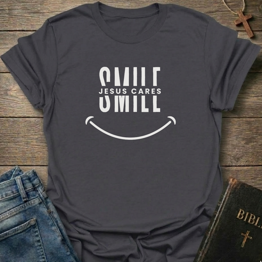 A dark charcoal gray graphic t-shirt with a white smiley face design and the text 'SMILE JESUS CARES' and 'SMILL' printed on the front. The shirt is styled in a flat-lay arrangement on a rustic wooden background, accompanied by blue jeans, a brown cross necklace, and a Bible.