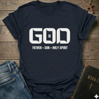 A navy short-sleeve graphic T-shirt laid flat on a rustic wooden surface, styled with blue jeans, a Bible, and a cross. The shirt has large white text reading 'GOD IS' with 'FATHER • SON • HOLY SPIRIT' underneath.