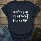 Navy blue t-shirt with 'Walking in Obedience brings life' text and cross graphic Christian script faith apparel conversation starter on a wooden surface.