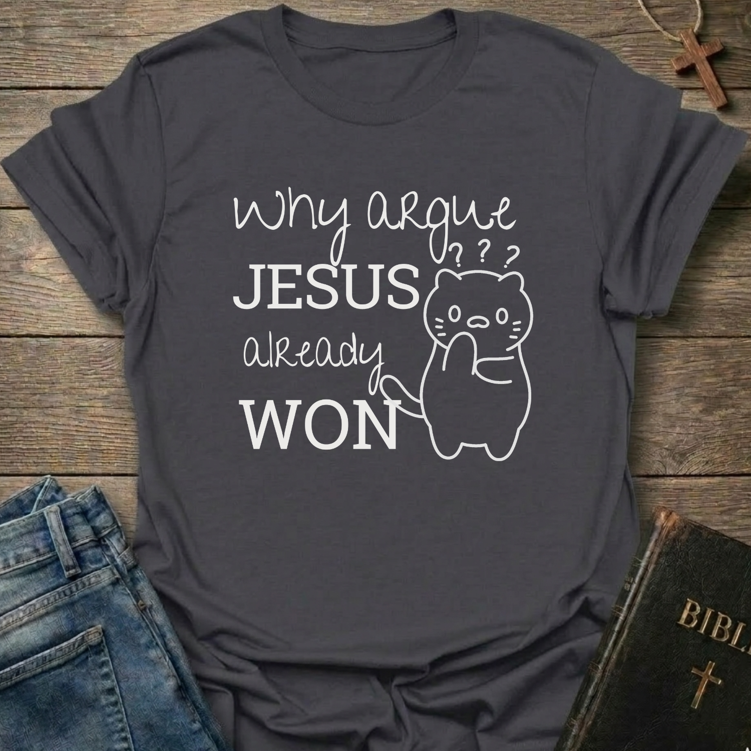 A dark charcoal gray graphic t-shirt laid flat on a rustic wooden background, styled with blue jeans, a Bible, and a wooden cross. The t-shirt features a white printed graphic that reads 'Why argue JESUS already WON' alongside a simple line drawing of a confused cat.