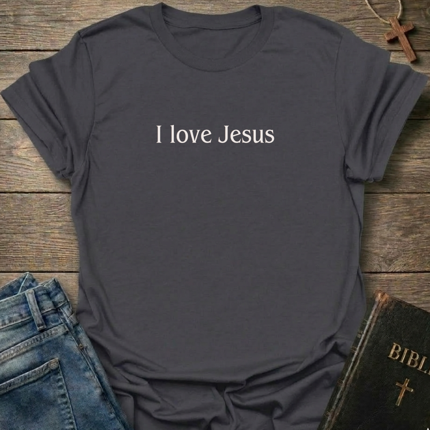 A dark charcoal gray t-shirt with the text 'I love Jesus' printed in white across the chest, styled in a flat-lay arrangement on a rustic wooden background with blue jeans and a Bible nearby.