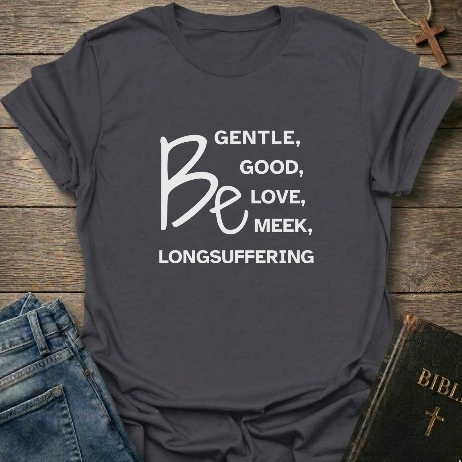 A dark charcoal gray graphic t-shirt with white text reading 'BE GENTLE, GOOD, LOVE, MEEK, LONGSUFFERING Be' laid flat on a rustic wooden background, styled with blue jeans and a black Bible.
