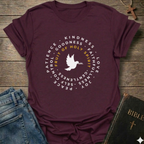A maroon short-sleeve graphic T-shirt laid flat on a rustic wooden surface, styled with blue jeans and a Bible. The shirt has a circular white and yellow print featuring a dove and the words 'PATIENCE', 'KINDNESS', 'GOODNESS', 'FAITH', 'LOVE', 'JOY', 'PEACE', and 'GENTLENESS', with 'FRUIT OF HOLY SPIRIT' in yellow.