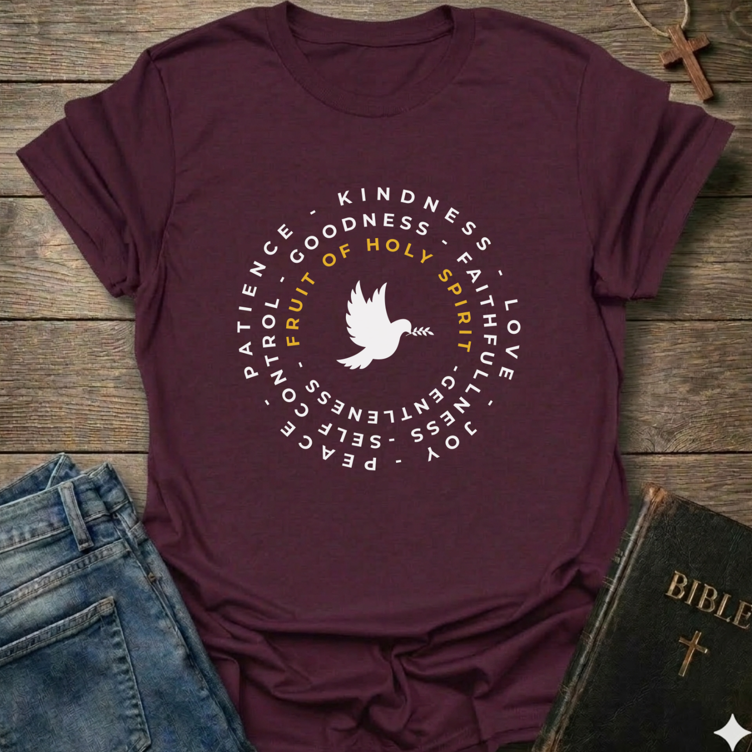A maroon short-sleeve graphic T-shirt laid flat on a rustic wooden surface, styled with blue jeans and a Bible. The shirt has a circular white and yellow print featuring a dove and the words 'PATIENCE', 'KINDNESS', 'GOODNESS', 'FAITH', 'LOVE', 'JOY', 'PEACE', and 'GENTLENESS', with 'FRUIT OF HOLY SPIRIT' in yellow.