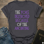Gray t-shirt with motivational text 'The yoke destroyed because of the anointing' Isaiah 10:27 faith apparel conversation starter on a wooden surface with jeans and a Bible.