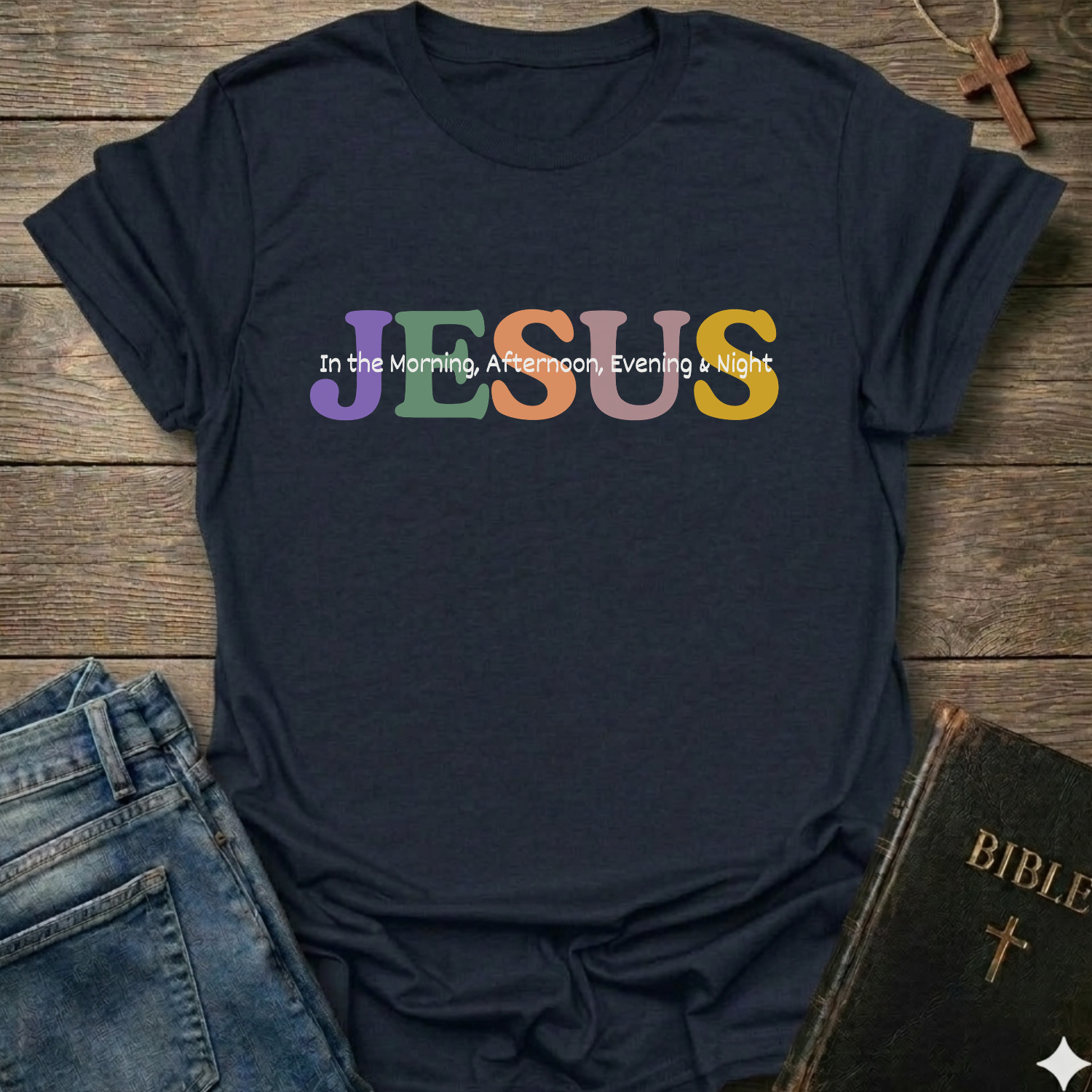 A navy blue graphic t-shirt laid flat on a rustic wooden background, styled with blue jeans, a Bible, and a cross necklace. The shirt features the word 'JESUS' in large, colorful pastel letters (purple, green, orange, yellow) with the phrase 'In the Morning, Afternoon, Evening & Night' printed in white script on top of it.